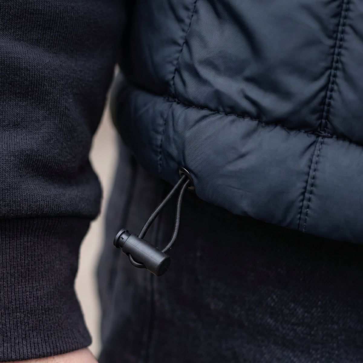 Close-up of a person wearing a dark navy quilted puffer jacket over a black ribbed sweater, showing the drawstring and black toggle.