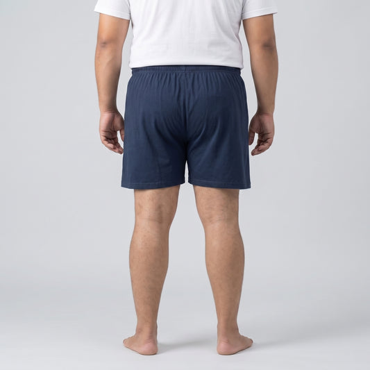A man viewed from the back wearing navy blue shorts and a white t-shirt, standing barefoot against a light gray studio background.