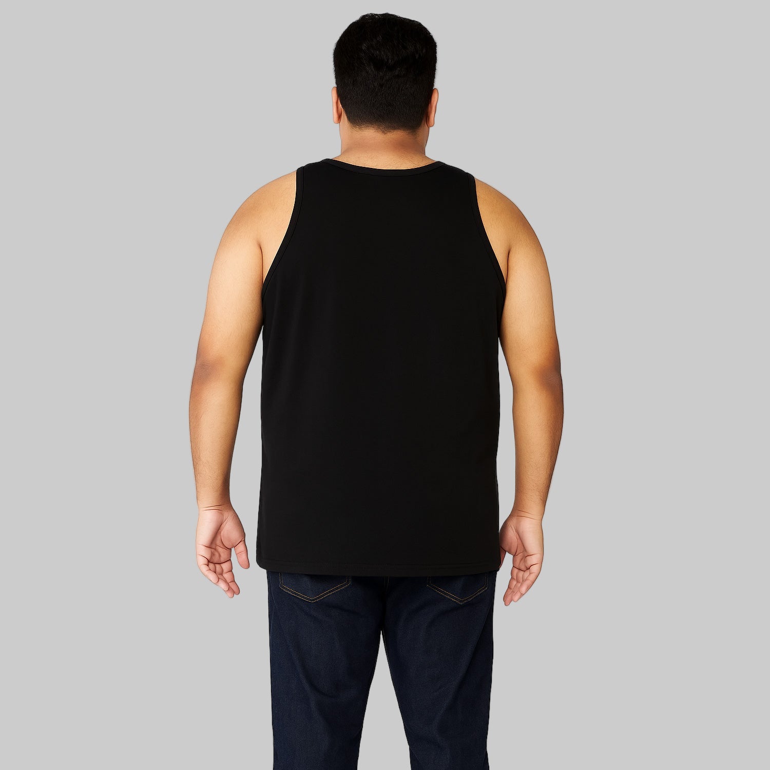 Plus-size Pakistani male model wearing a black sleeveless cotton tank top with dark jeans on a light-gray background, relaxed fit casual wear.
