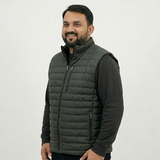A man wearing a dark gray quilted puffer vest over a black long-sleeve shirt, standing against a plain light gray background.