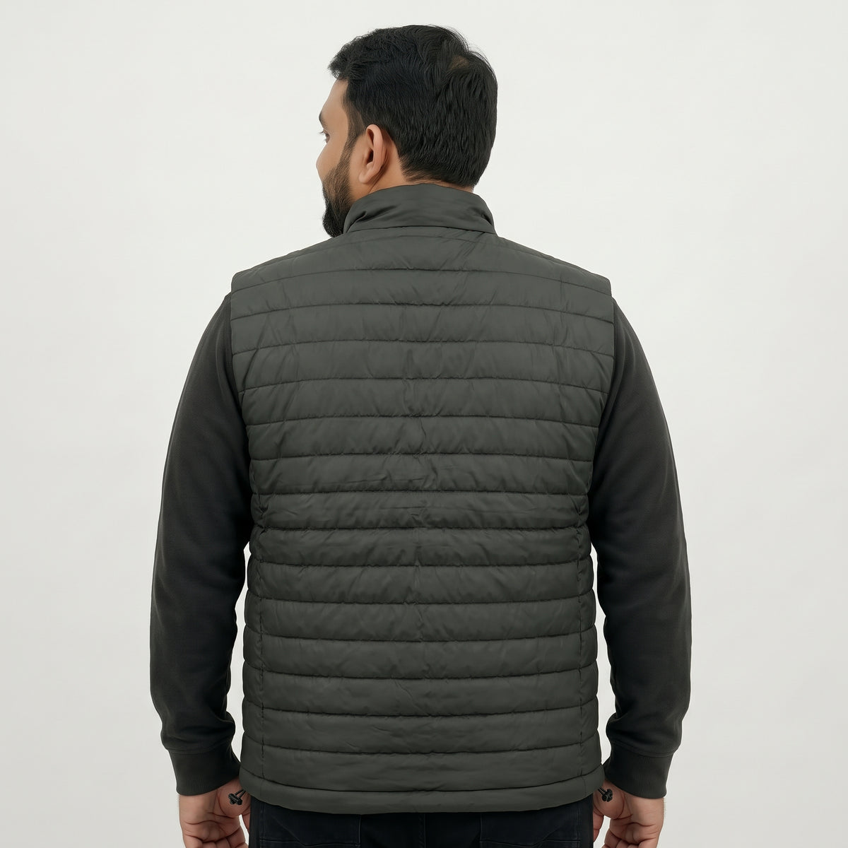 A man viewed from the back wearing a dark gray quilted puffer vest over a black long-sleeve shirt, against a plain light gray studio background.