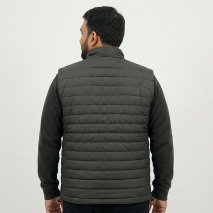 A man viewed from the back wearing a dark gray quilted puffer vest over a black long-sleeve shirt, against a plain light gray studio background.
