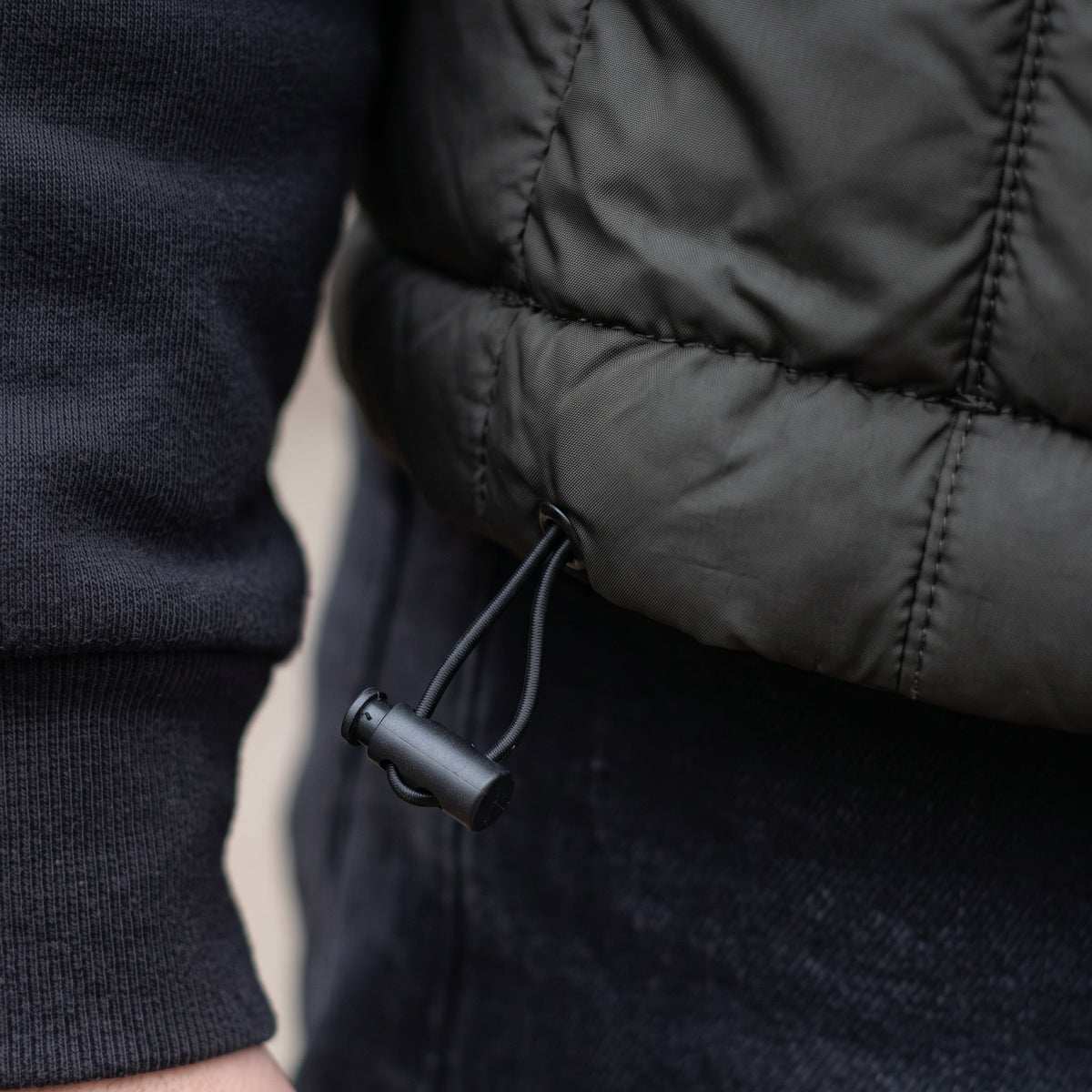 Close-up of a black puffer jacket with a drawstring hem, worn over a dark ribbed sweater and dark pants.