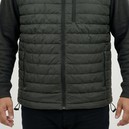 A close-up of a person wearing a dark olive green quilted puffer vest over a black long-sleeve shirt, showing the front zipper, chest pocket, and side zip pockets.