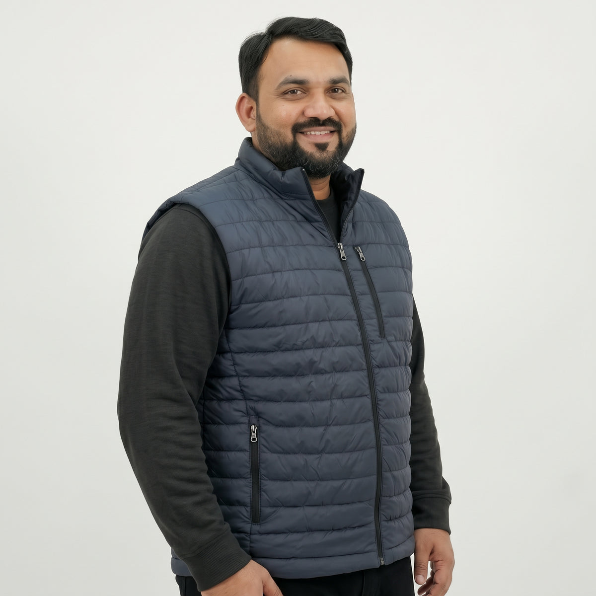 A man wearing a navy blue quilted puffer vest over a black long-sleeve shirt, photographed against a plain light gray studio background.