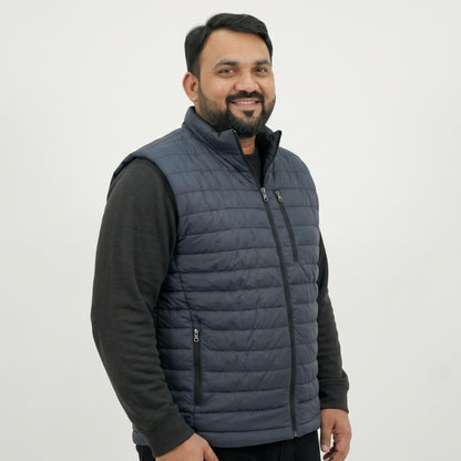 A man wearing a navy blue quilted puffer vest over a black long-sleeve shirt, photographed against a plain light gray studio background.