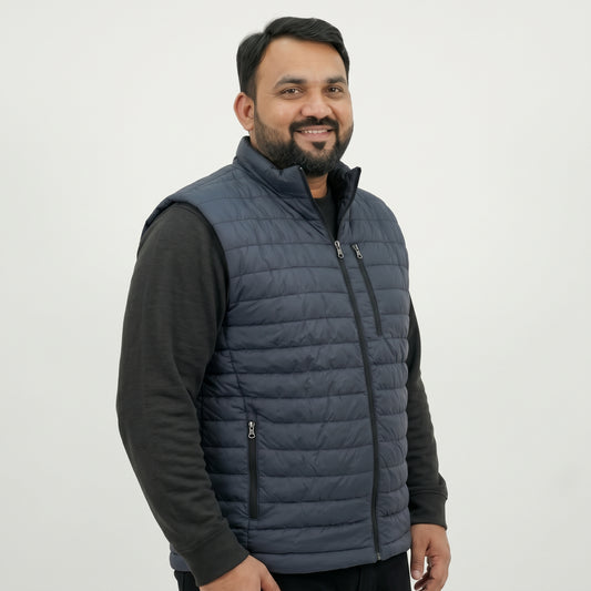 A man wearing a navy blue quilted puffer vest over a black long-sleeve shirt, photographed against a plain light gray studio background.