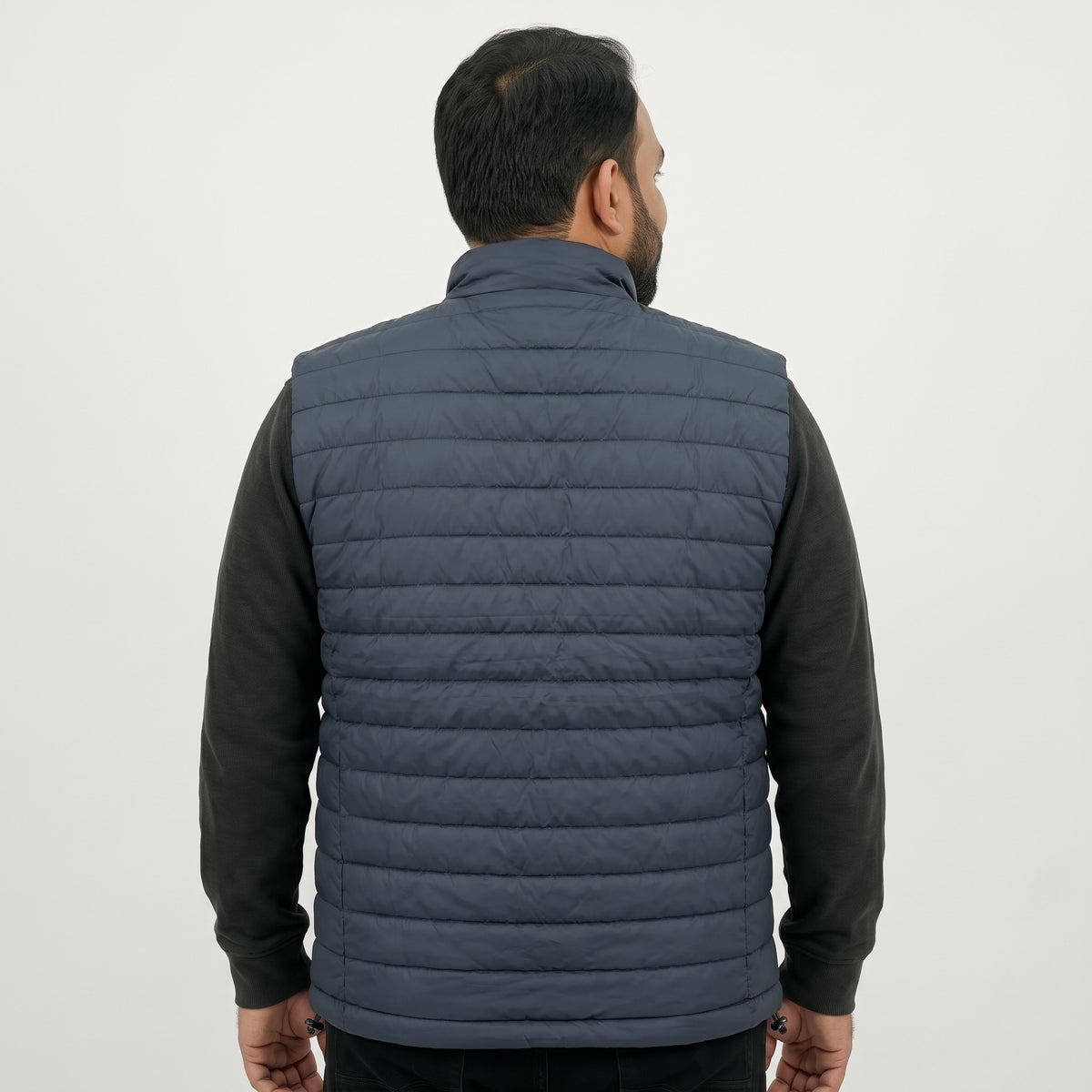 A man viewed from the back wearing a dark slate blue quilted puffer vest over a black long-sleeve shirt, against a plain light gray background.