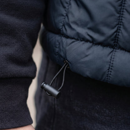 Close-up of a person wearing a dark navy quilted puffer jacket over a black ribbed sweater, showing the drawstring and black toggle.