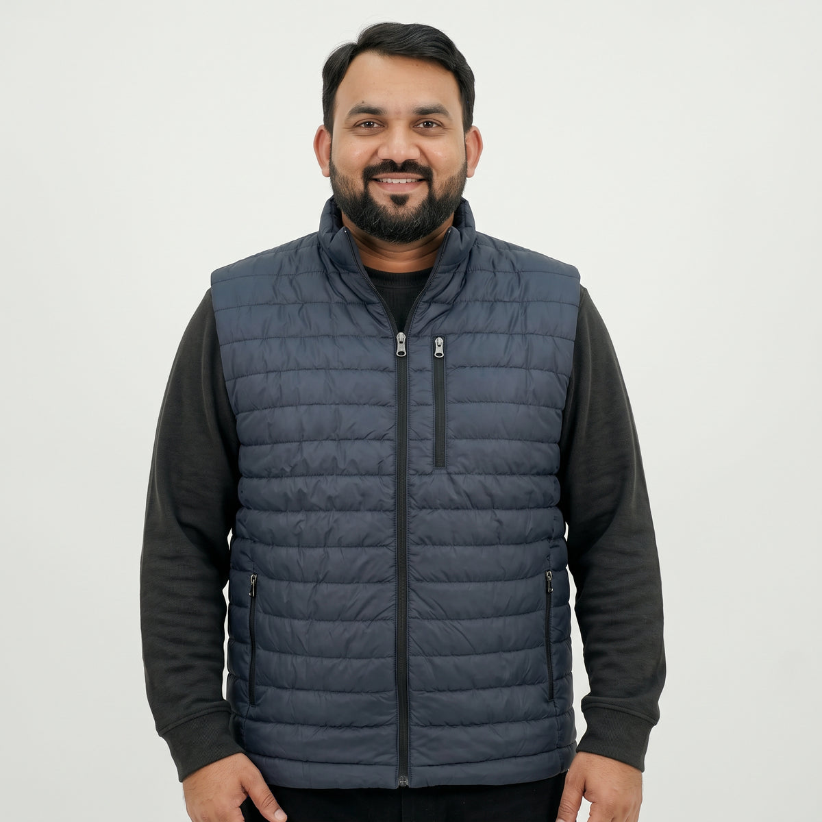 A man wearing a navy blue quilted zip-up vest over a black long-sleeve shirt, standing against a plain light gray background.