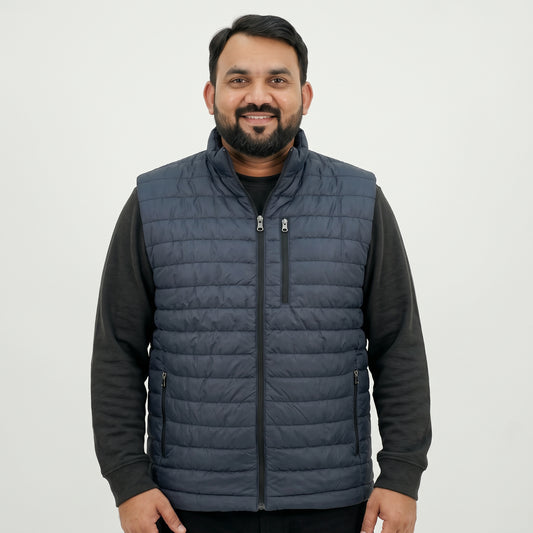 A man wearing a navy blue quilted zip-up vest over a black long-sleeve shirt, standing against a plain light gray background.