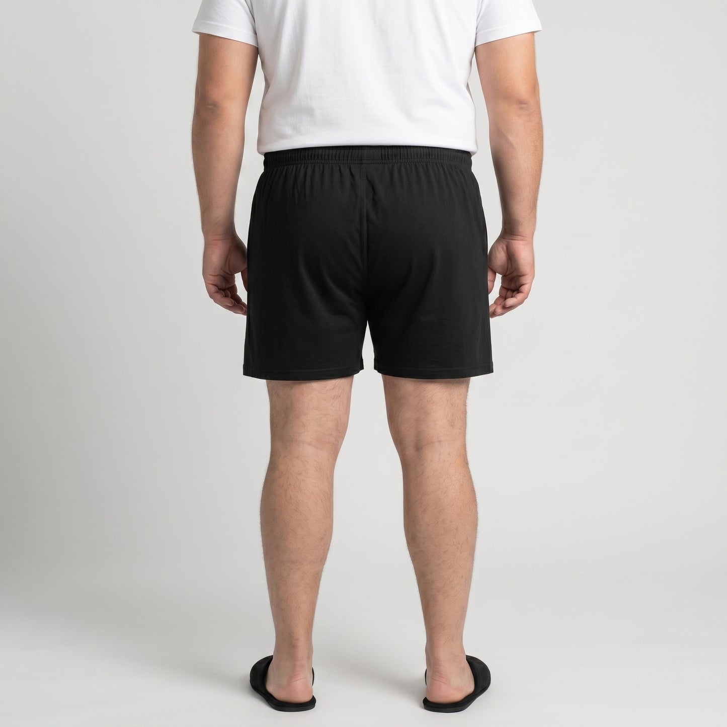 A man viewed from behind wearing black boxer shorts, a white t-shirt, and black slide sandals, standing against a light gray studio background.