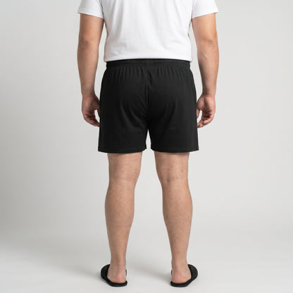 A man viewed from behind wearing black boxer shorts, a white t-shirt, and black slide sandals, standing against a light gray studio background.