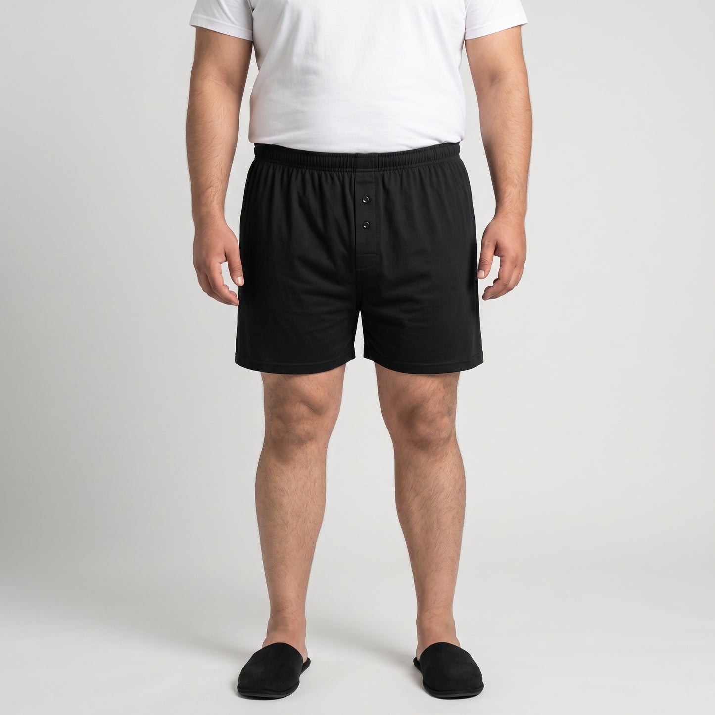 A man wearing black boxer briefs, a white t-shirt, and black slippers, standing against a light gray studio background.