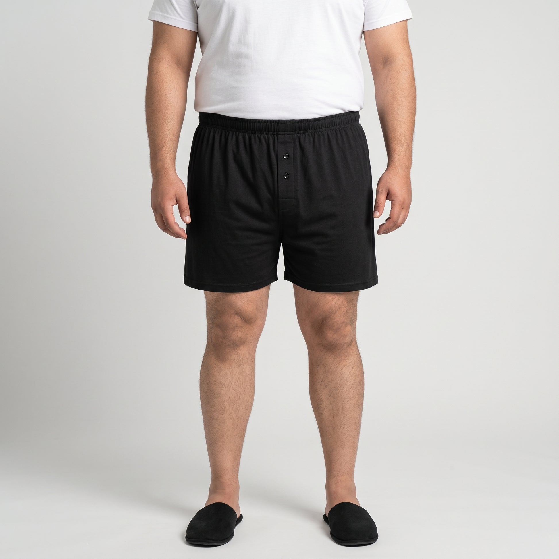 A man wearing black boxer briefs, a white t-shirt, and black slippers, standing against a light gray studio background.