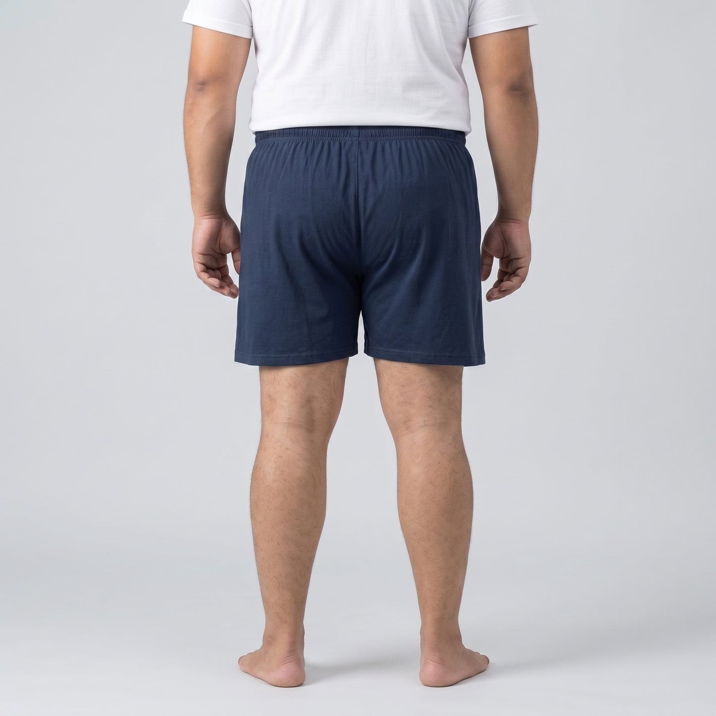 A man viewed from the back wearing navy blue shorts and a white t-shirt, standing barefoot against a light gray studio background.