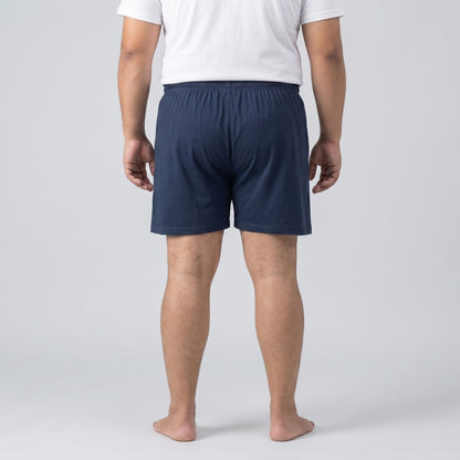 A man viewed from the back wearing navy blue shorts and a white t-shirt, standing barefoot against a light gray studio background.
