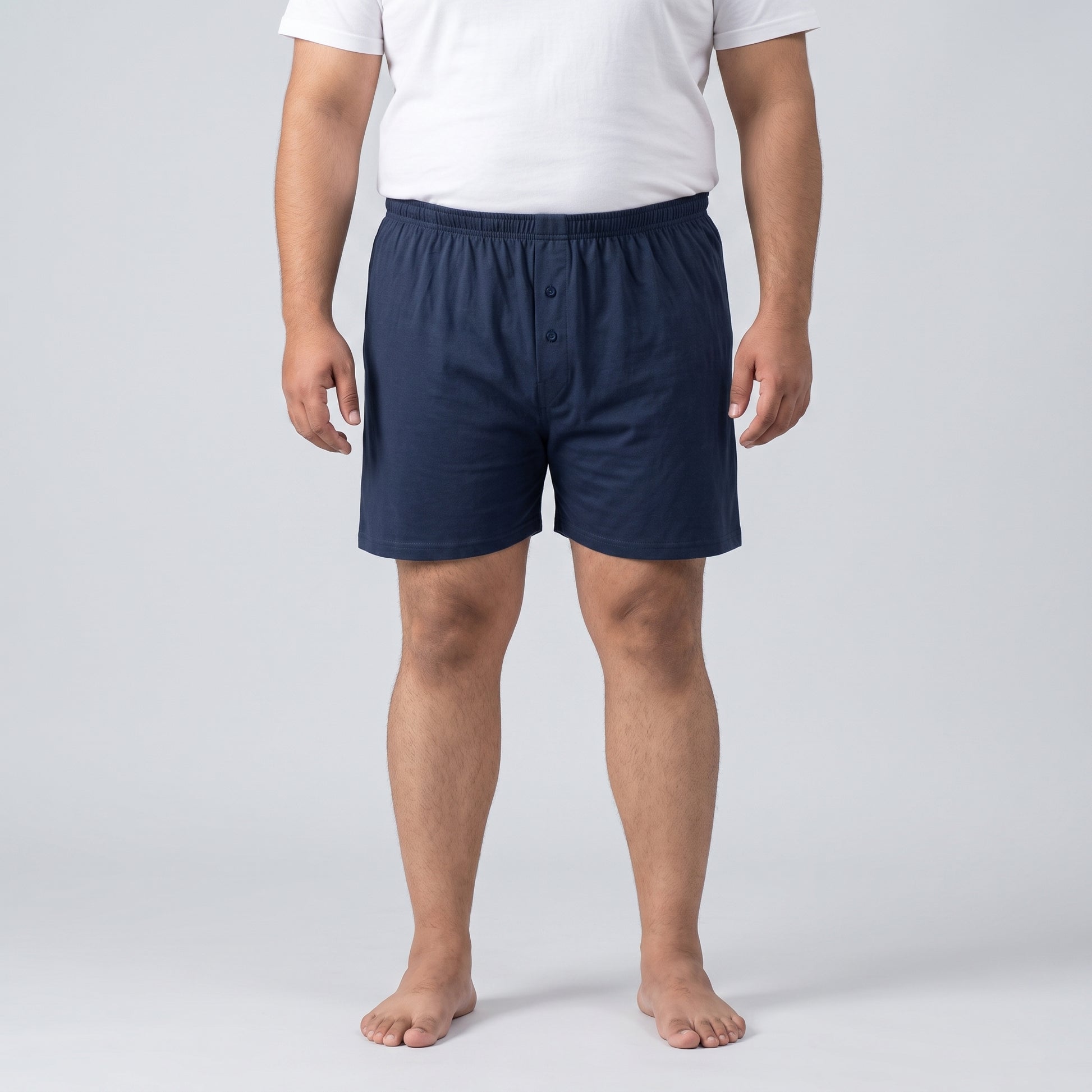 A man wearing navy blue boxer shorts and a white t-shirt, standing against a light gray studio background.