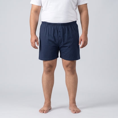 A man wearing navy blue boxer shorts and a white t-shirt, standing against a light gray studio background.