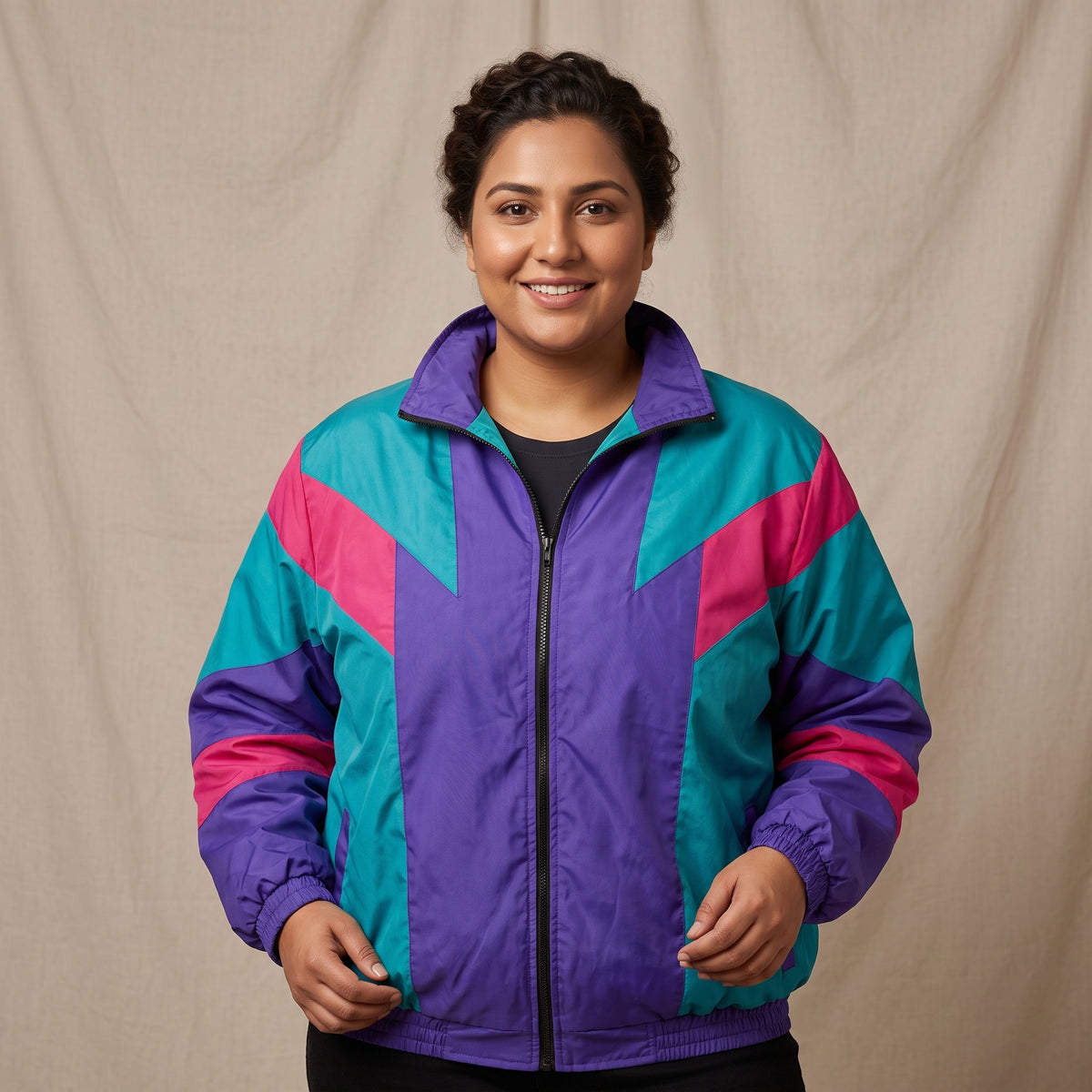 A woman wearing a retro 90s-style windbreaker jacket with geometric color-blocked panels in purple, teal, and hot pink, standing against a neutral beige fabric backdrop.
