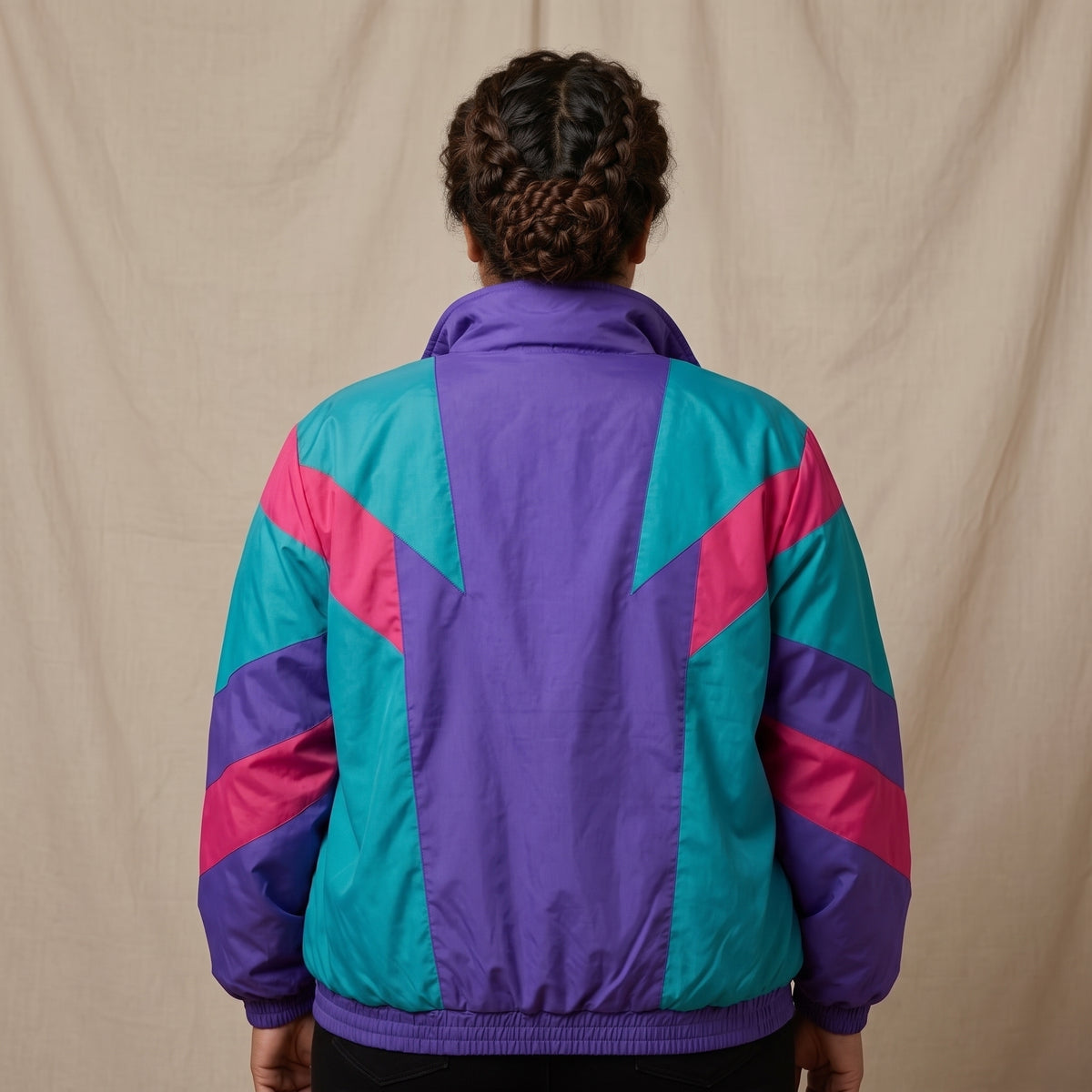 A person viewed from behind wearing a retro-style windbreaker jacket with a purple back panel and geometric teal and pink panels on the shoulders and sleeves, posed against a neutral beige fabric backdrop.