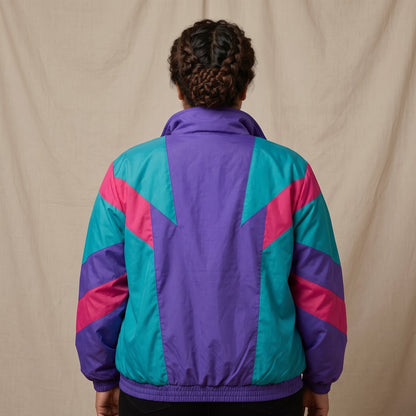 A person viewed from behind wearing a retro-style windbreaker jacket with a purple back panel and geometric teal and pink panels on the shoulders and sleeves, posed against a neutral beige fabric backdrop.
