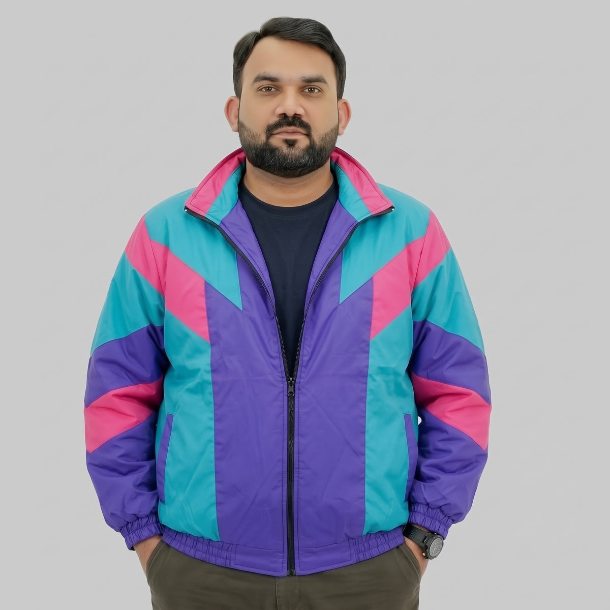 A man wearing a colorful geometric windbreaker jacket with purple, teal, and pink panels, standing against a solid gray studio background.